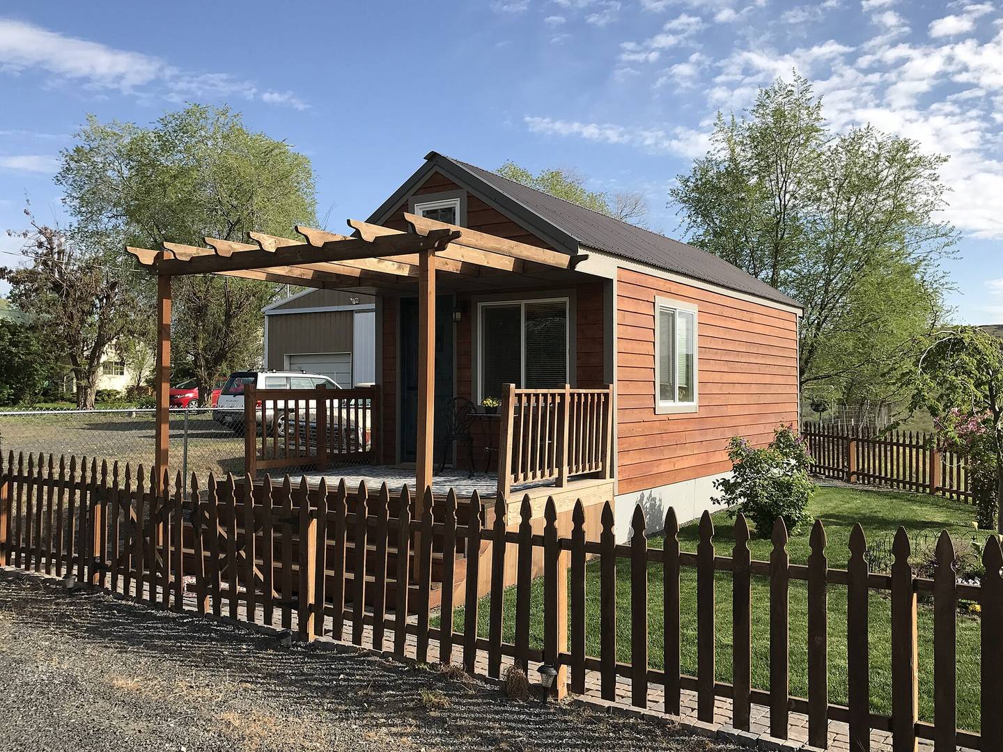 A detached backyard cottage ADU with wooden cladding, a pergola terrace and a picket fence