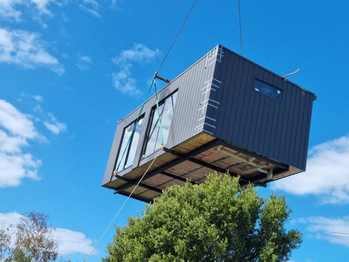 A prefabricated building module with large windows being lifted by a crane against a blue sky