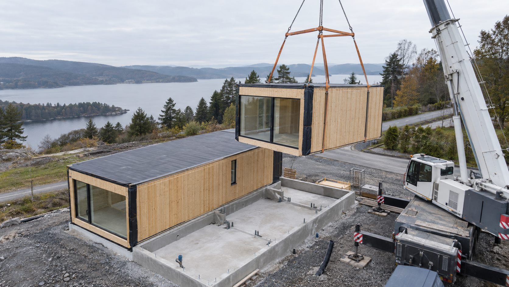 Editorial visual of a timber volumetric module being lifted onto prepared concrete foundations beside an installed module