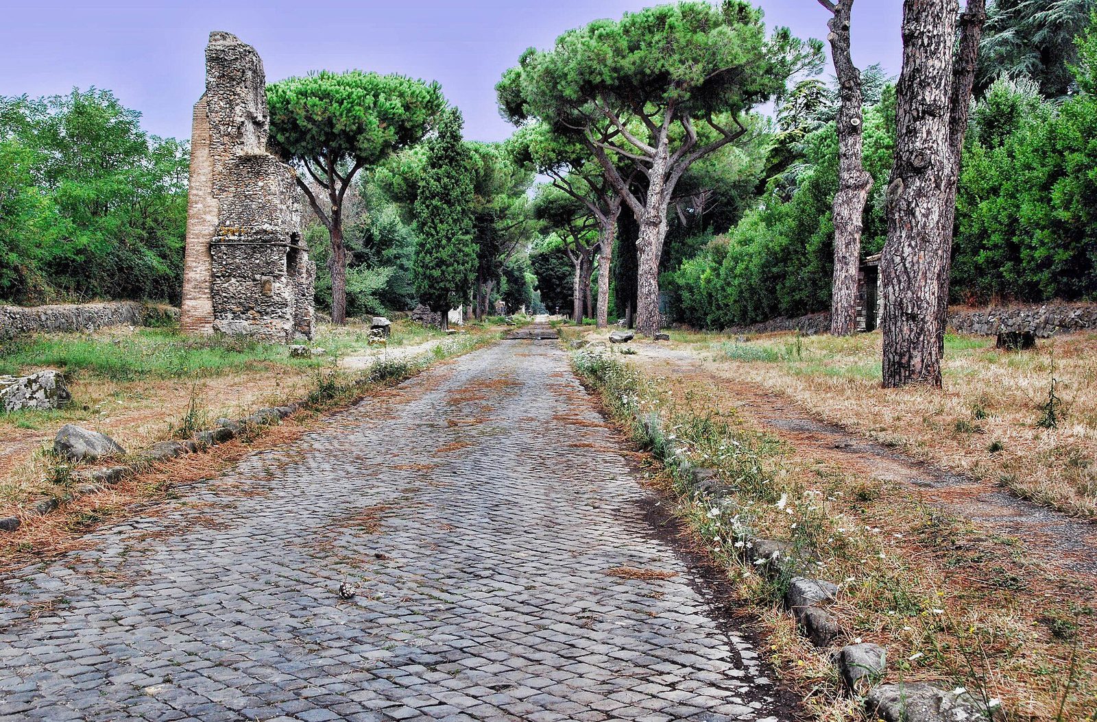 The basalt-paved Via Appia Antica outside Rome, flanked by umbrella pines and the broken remains of a Roman tomb
