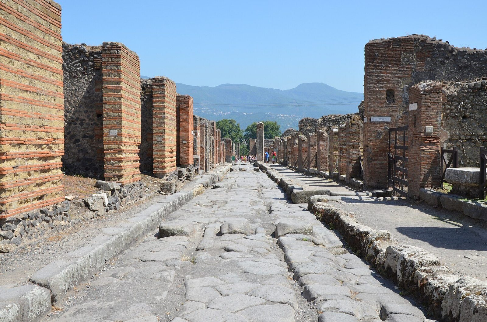 A paved street at Pompeii: worn stone slabs and raised pavements running between brick house fronts, with Vesuvius on the horizon