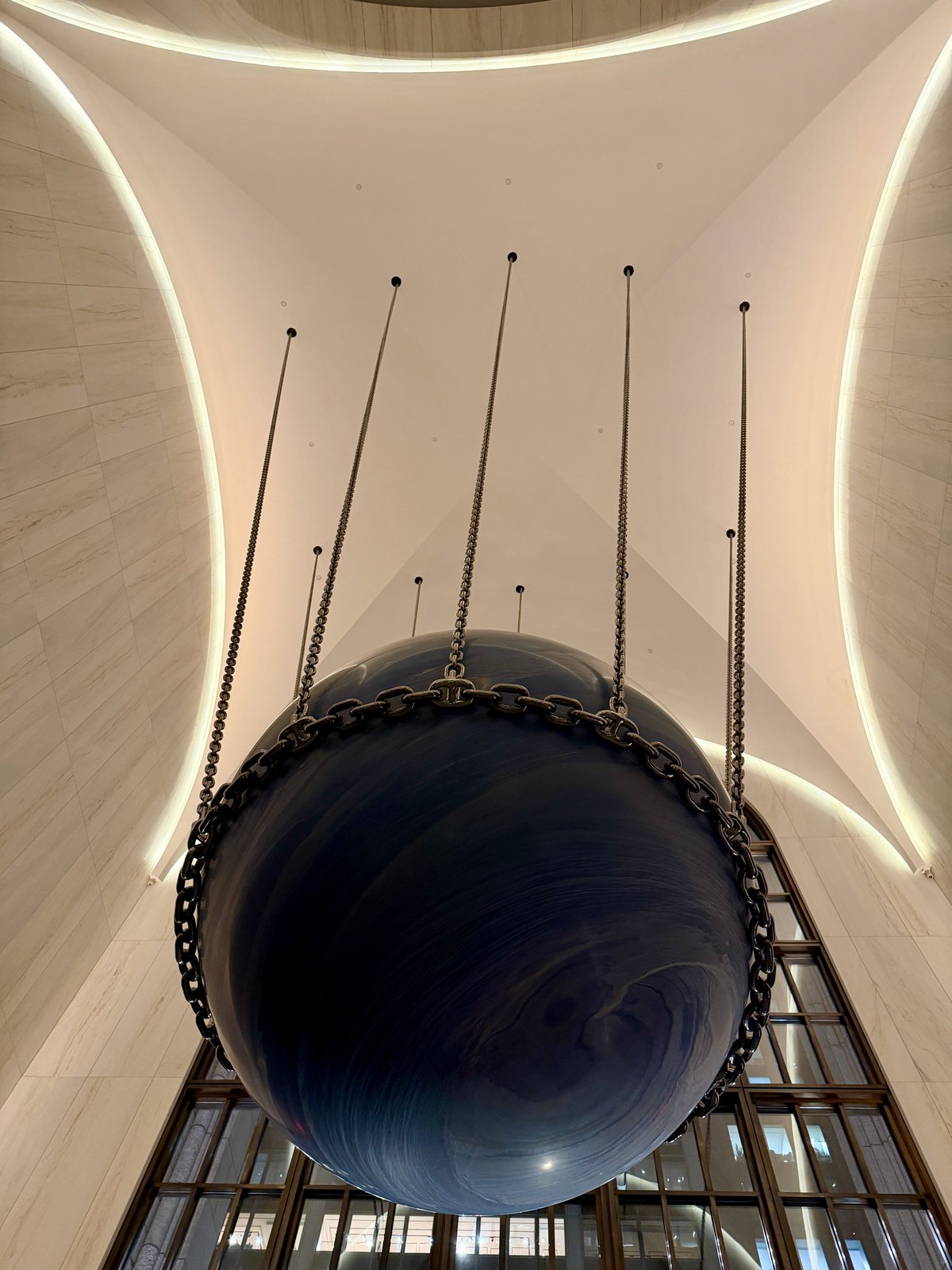 The Solid Sky sculpture hanging in the atrium of 550 Madison Avenue, a dark sphere suspended beneath a vaulted ceiling.