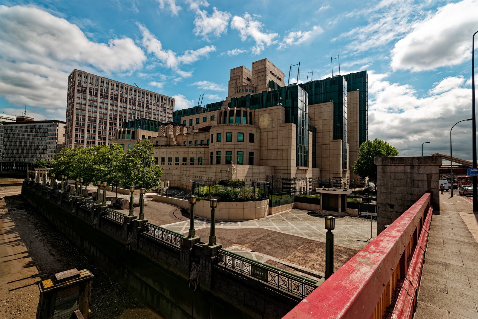 The MI6 (SIS) Building in London seen from the riverside walkway, its stepped stone and green-glass mass rising behind trees.