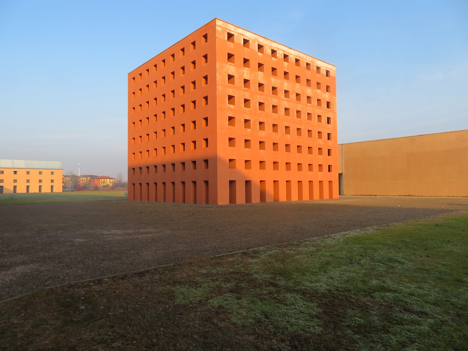 The red cubic ossuary block of San Cataldo Cemetery in Modena, its grid of square openings rising over an empty forecourt.