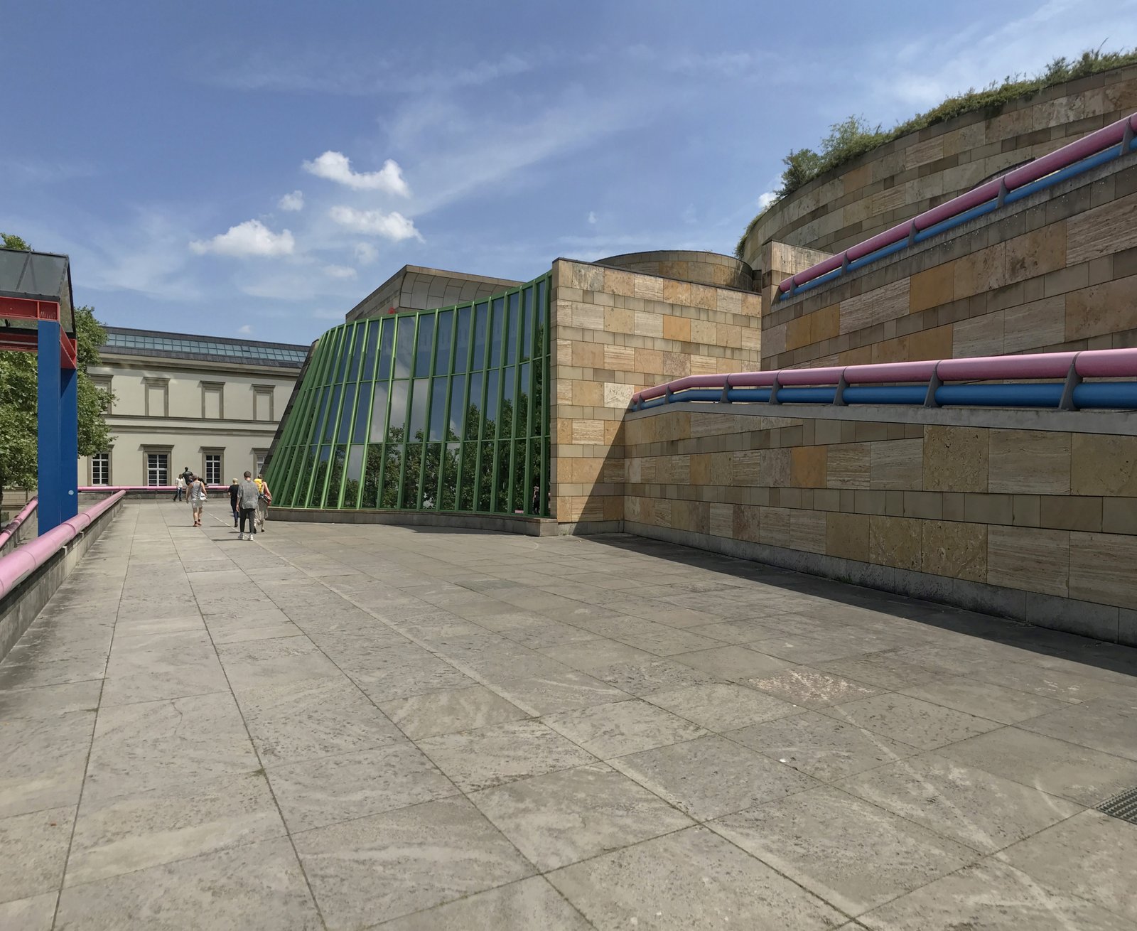 The Neue Staatsgalerie in Stuttgart seen from its paved terrace, with green glazed walls, stone ramps and pink and blue handrails.