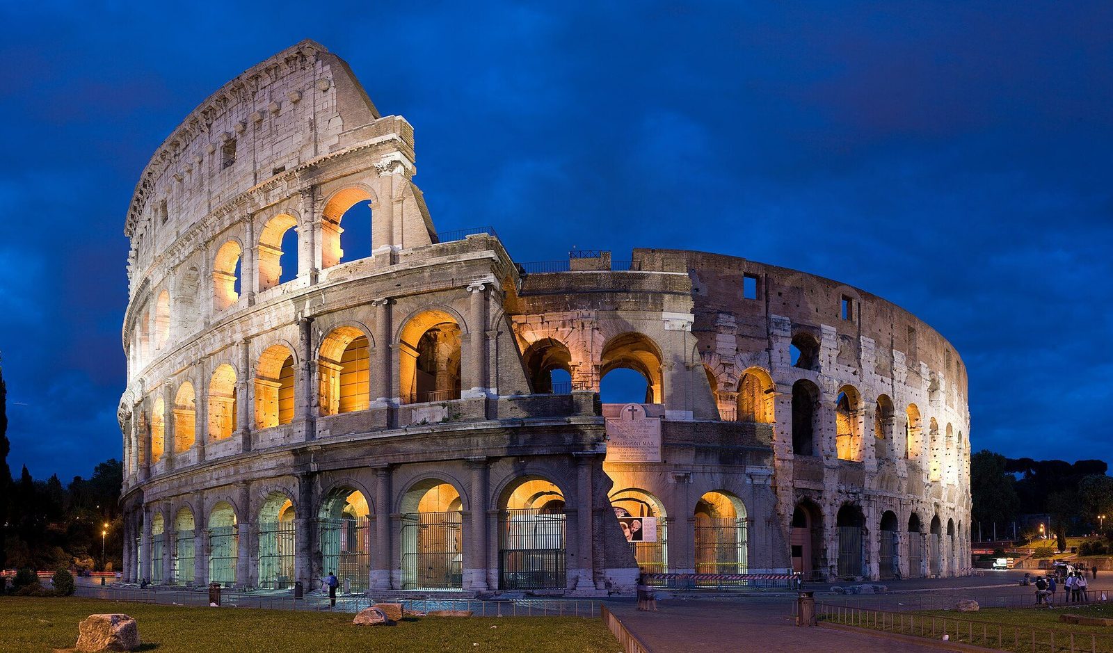 The Colosseum in Rome at dusk: its three tiers of travertine arches lit from within against a deep blue evening sky